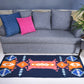 A gray couch adorned with two decorative pillows—one blue and one pink—sits in a stylish interior. In front of the couch, a colorful patterned rug features shades of orange, blue, and white. A small side table next to the couch holds a hat and some magazines, while a green plant adds a touch of nature to the scene.
