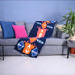 A cozy gray sofa adorned with colorful cushions in blue and pink. A vibrant patterned throw blanket, featuring bold orange and blue geometric designs, drapes over the arm of the sofa. A green potted plant is visible in the corner, enhancing the inviting atmosphere of the living space.