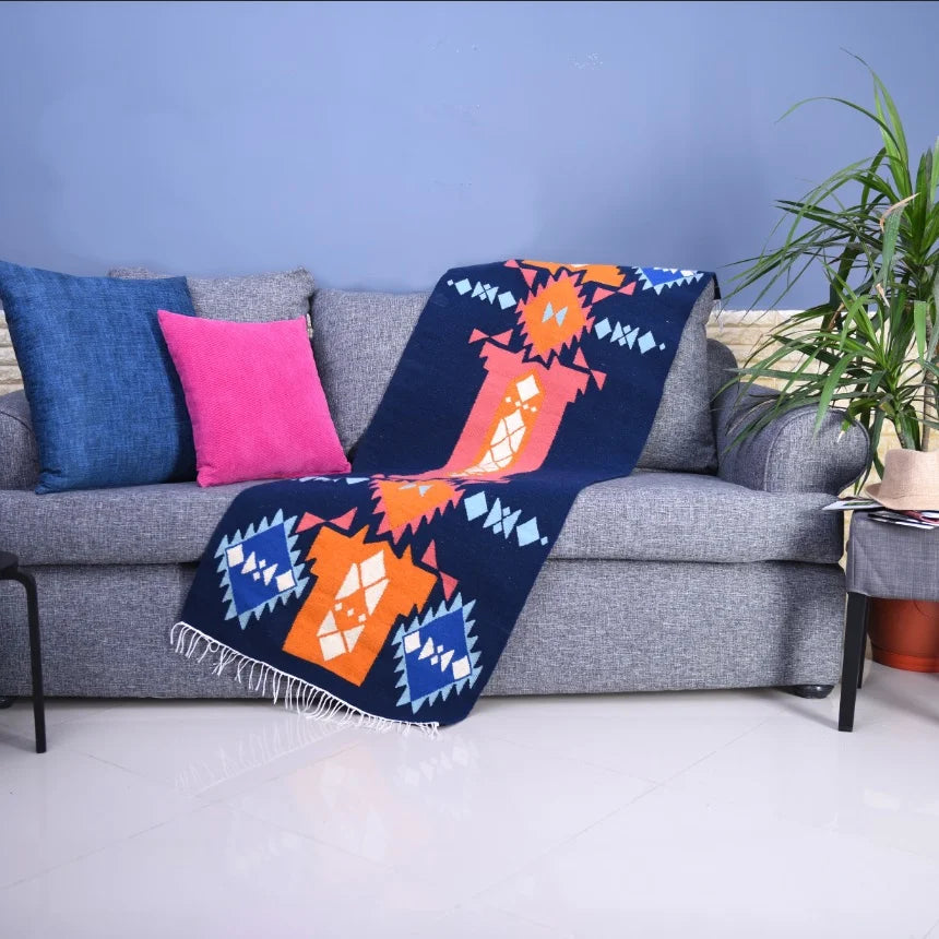 A cozy gray sofa adorned with colorful cushions in blue and pink. A vibrant patterned throw blanket, featuring bold orange and blue geometric designs, drapes over the arm of the sofa. A green potted plant is visible in the corner, enhancing the inviting atmosphere of the living space.