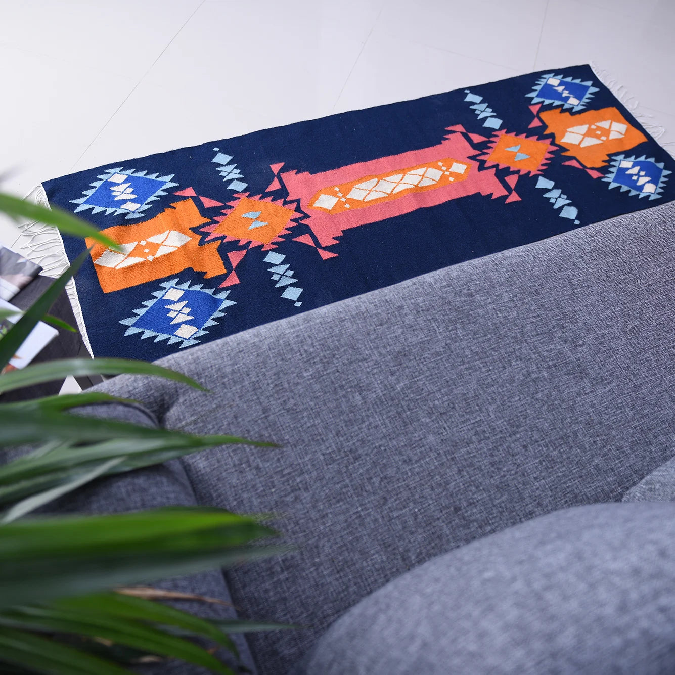 A colorful patterned rug in vibrant shades of orange, blue, and white, placed on a light-colored floor. In the foreground, a gray couch is partially visible, along with a green plant, creating a cozy interior setting.