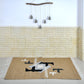A woven rug displayed on a light-colored tiled floor, featuring a geometric design with shades of beige, black, gray, and white. Above the rug, a decorative wall hanging made of wood and tassels complements the earthy tones of the space. The textured stone wall behind adds warmth to the overall aesthetic.