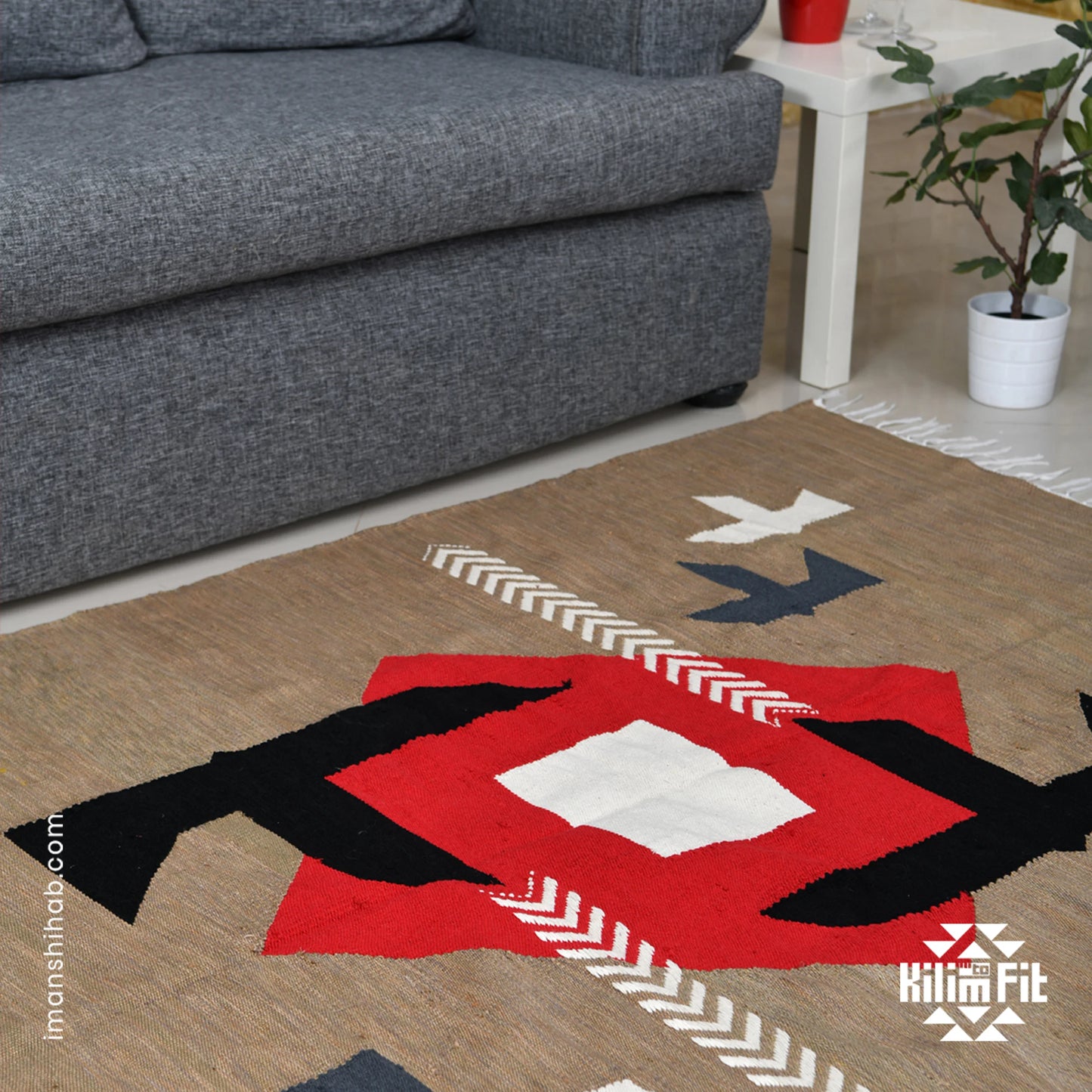 A close-up of a woven rug featuring a striking geometric pattern with a central red diamond, complemented by black, white, and gray shapes, including arrows and stylized birds. In the background, a gray couch is visible, along with a small white side table and a green plant, creating a cozy and modern living space.