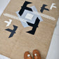 A woven rug featuring a geometric design in earthy tones, with a central light gray shape surrounded by black and white accents, including stylized birds. In the foreground, a pair of brown sandals with a decorative buckle rests on the rug, adding a casual and inviting touch to the scene. The light-colored tiled floor enhances the rug's warm colors.
