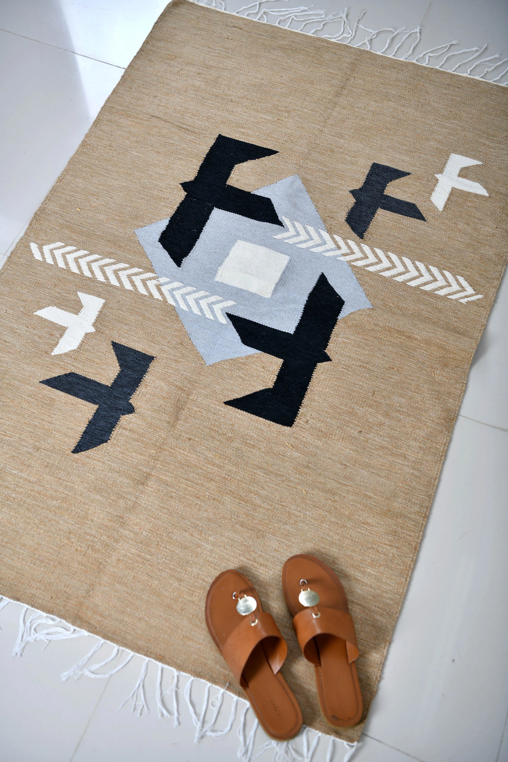 A woven rug featuring a geometric design in earthy tones, with a central light gray shape surrounded by black and white accents, including stylized birds. In the foreground, a pair of brown sandals with a decorative buckle rests on the rug, adding a casual and inviting touch to the scene. The light-colored tiled floor enhances the rug's warm colors.