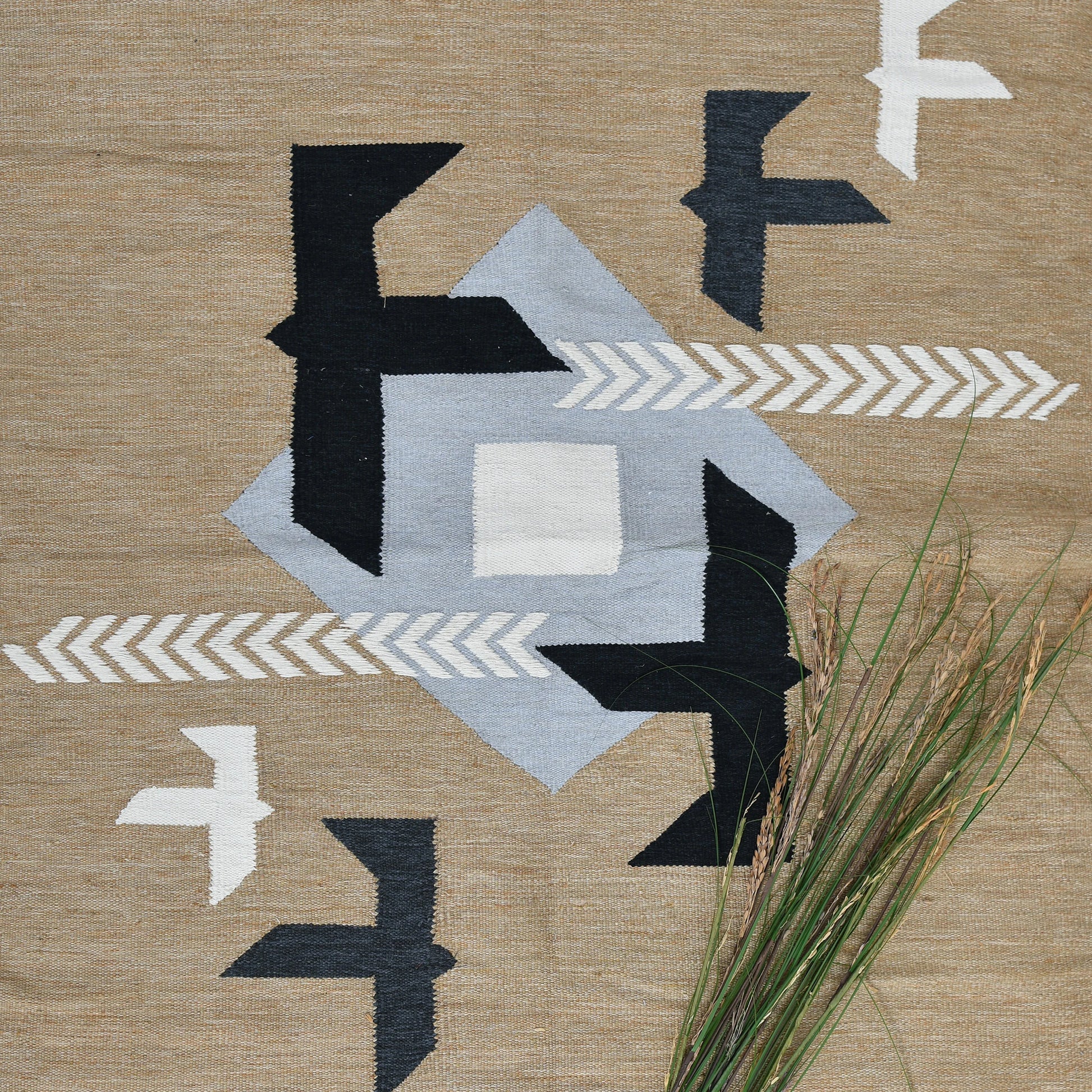 A close-up of a woven rug featuring a geometric design in neutral tones, with a central light gray shape and surrounding black and white elements, including stylized birds and arrows. In the foreground, a few strands of grass add a natural touch, enhancing the earthy aesthetic of the rug. The texture of the rug is highlighted against the light background.