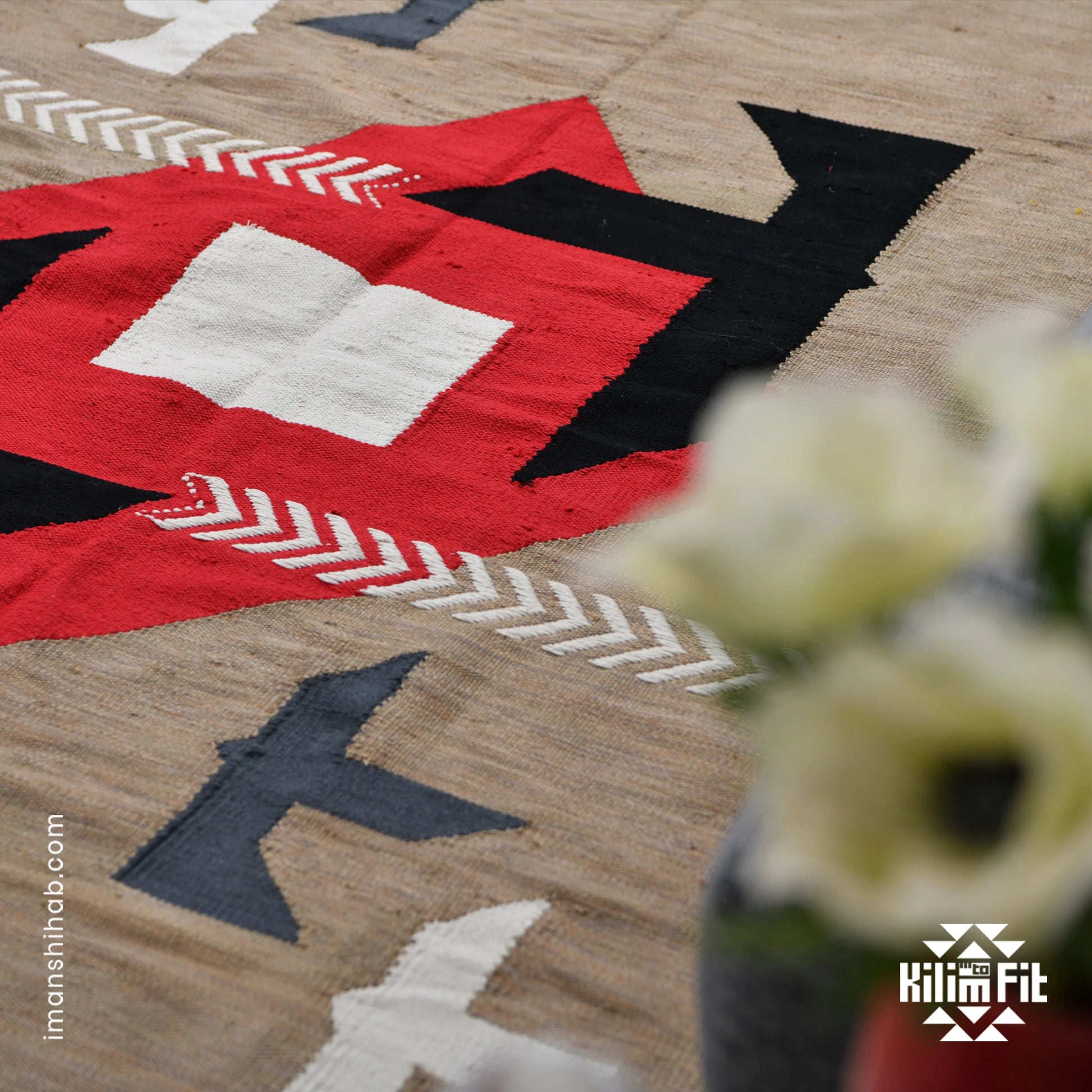 A close-up of a woven rug showcasing a vibrant geometric design with a central red diamond, accented by black, white, and gray shapes, including arrows and stylized birds. In the foreground, blurred white flowers in a vase add a soft touch, while the rug's intricate patterns remain the focal point of the image.