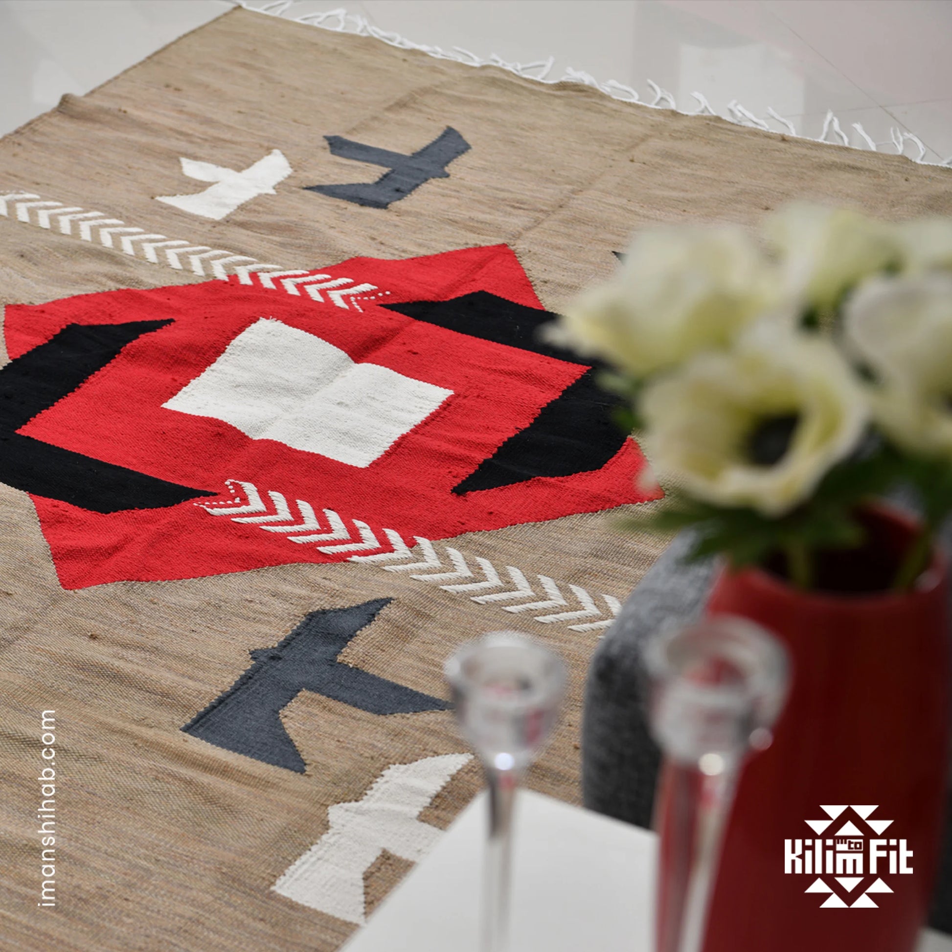 A close-up view of a woven rug featuring a bold geometric design with a central red diamond, surrounded by black, white, and gray shapes, including arrows and stylized birds. In the foreground, a decorative vase with white flowers adds a touch of elegance, and two glasses are partially visible. The setting is bright and modern, emphasizing the rug's vibrant colors and textures.