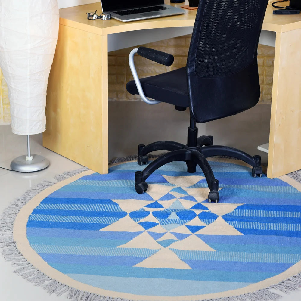 A round area rug with a geometric design in shades of blue and cream, featuring a star pattern at its center. The rug is positioned under a black office chair in a workspace with a light wooden desk and a desk lamp. The setting conveys a modern and organized office environment.