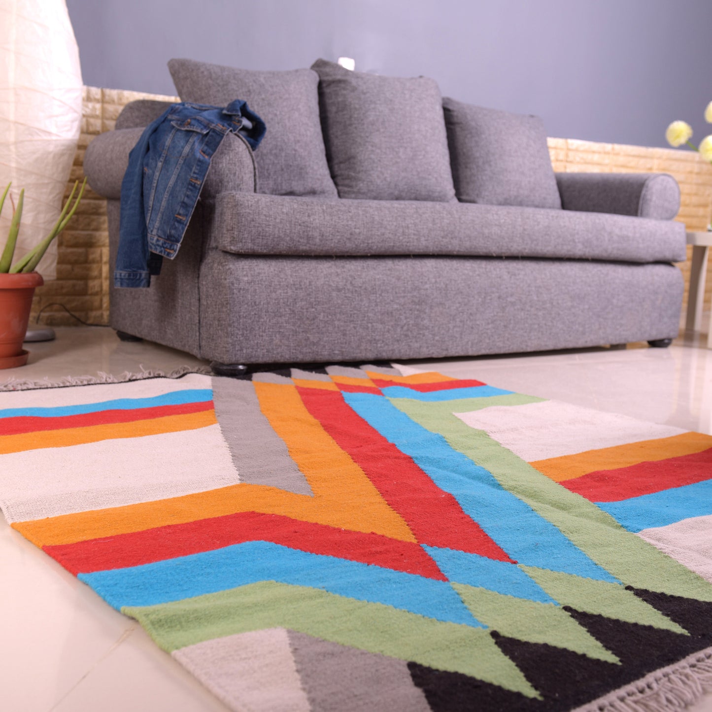 A vibrant, rectangular woven rug featuring a dynamic geometric pattern in bright colors, including turquoise, orange, red, and yellow, is placed on a light-colored tiled floor. In the background, a gray sofa with cushions and a denim jacket draped over the arm adds a cozy touch to the modern living space. Potted plants and soft lighting create an inviting atmosphere.