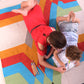 An overhead view of a young girl in a red dress and a small child in a light blue shirt lying on a colorful woven rug, engaged in coloring together. The rug features a vibrant geometric pattern with bold colors like turquoise, orange, and yellow. Colored pencils are scattered nearby, adding to the playful atmosphere. The gray sofa in the background completes the cozy scene.