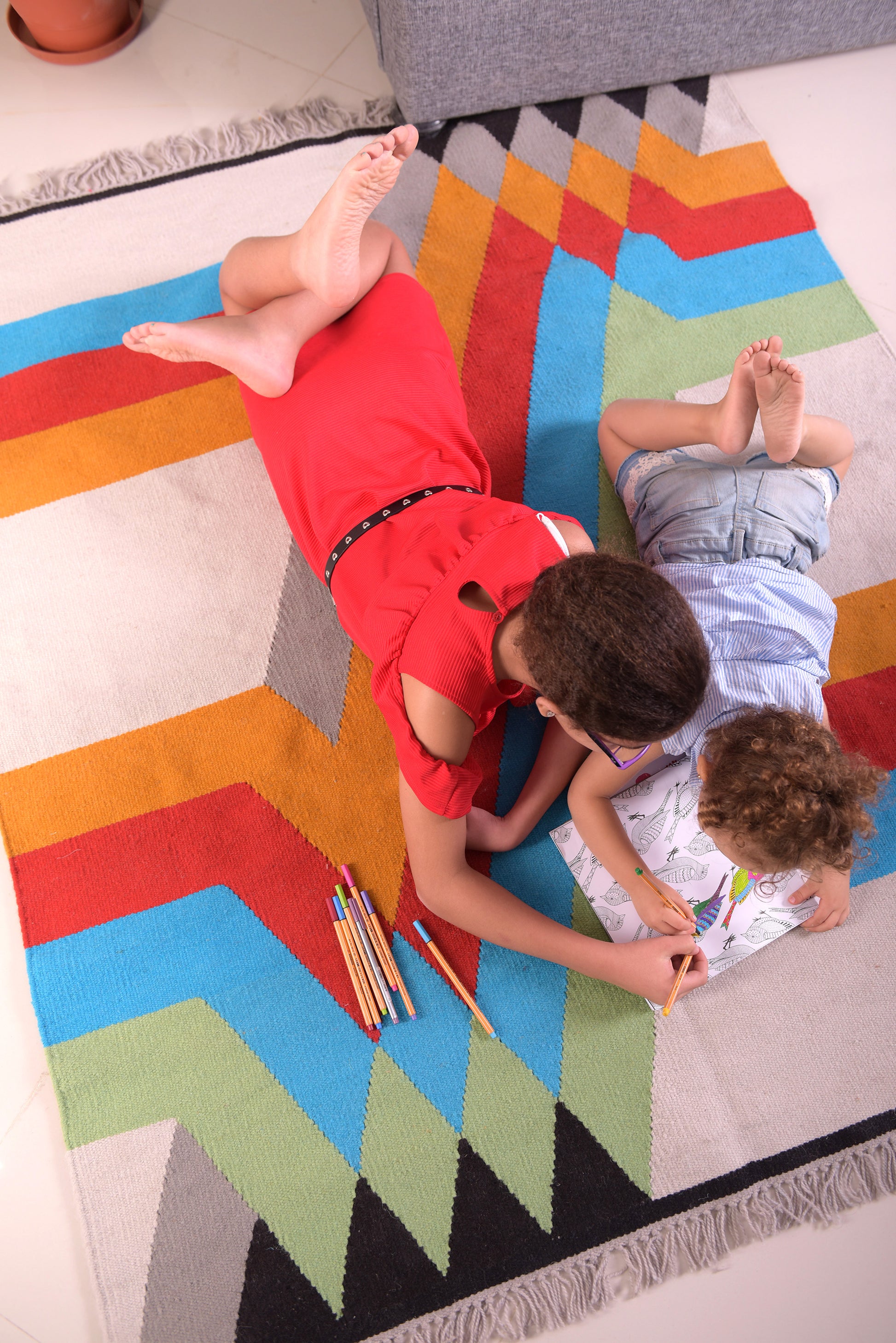 An overhead view of a young girl in a red dress and a small child in a light blue shirt lying on a colorful woven rug, engaged in coloring together. The rug features a vibrant geometric pattern with bold colors like turquoise, orange, and yellow. Colored pencils are scattered nearby, adding to the playful atmosphere. The gray sofa in the background completes the cozy scene.