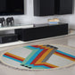 A round woven rug with a bold geometric design in vibrant colors, including turquoise, orange, red, and gray, is placed on a light-colored tiled floor. In the background, a modern black media console holds decorative elements, including two small elephant figurines and stacks of books. The contemporary setting enhances the rug's colorful pattern.