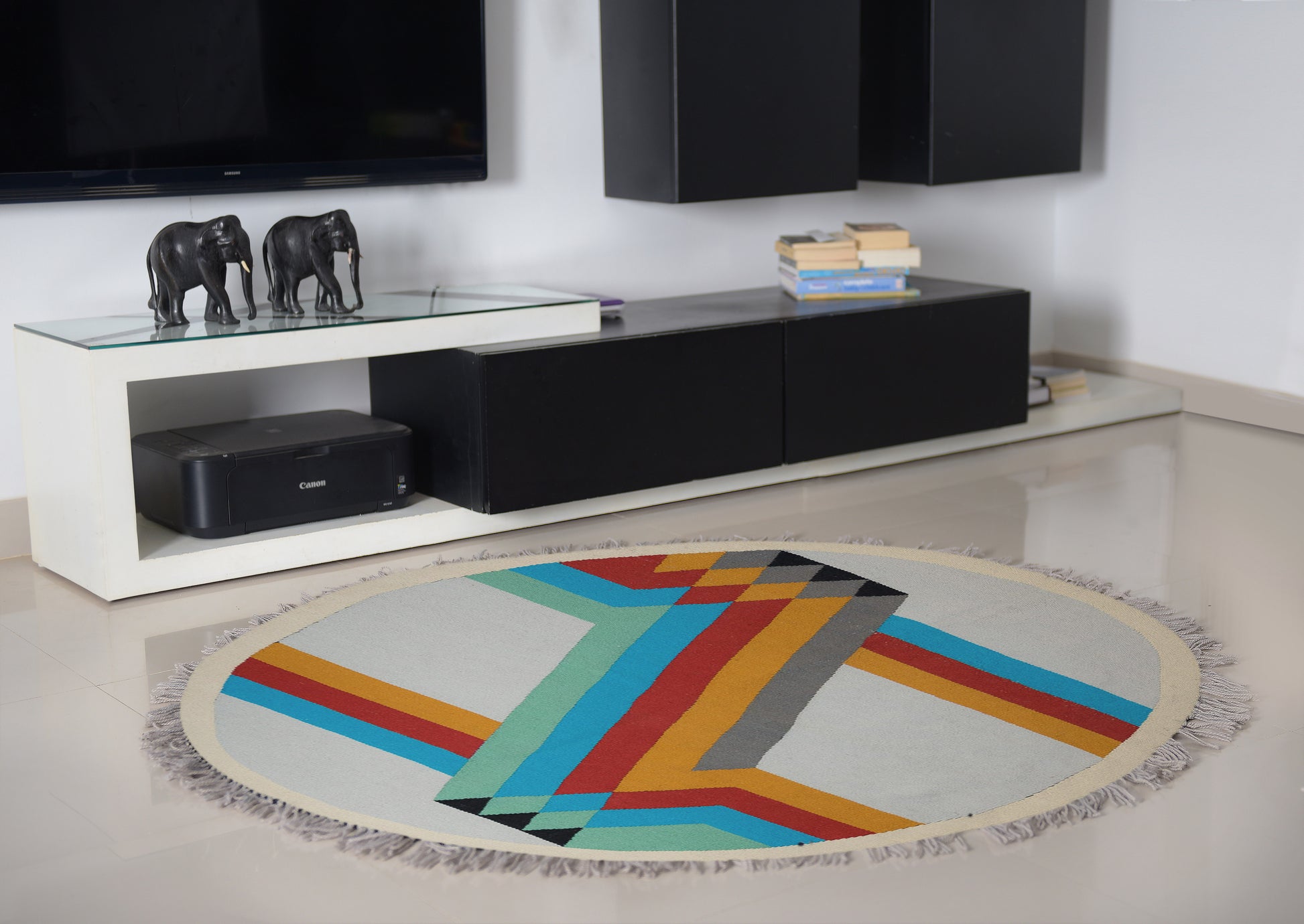 A round woven rug with a bold geometric design in vibrant colors, including turquoise, orange, red, and gray, is placed on a light-colored tiled floor. In the background, a modern black media console holds decorative elements, including two small elephant figurines and stacks of books. The contemporary setting enhances the rug's colorful pattern.