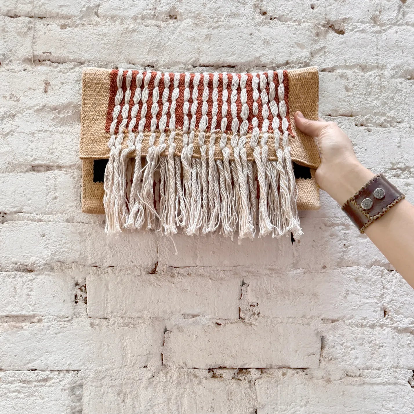 A woven clutch bag featuring a beige base with red and white vertical stripes and fringed detailing at the bottom.