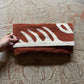 "A hand holding a terracotta woven clutch featuring abstract white leaf patterns, set against a textured stone surface adorned with ancient carvings. The image captures the clutch's design and the intricate background."