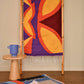 A colorful woven rug displayed on a wooden rack, featuring bold shapes in yellow, orange, and purple. Beside it, another smaller rug with a blue and black design is partially visible. A wooden cylindrical pedestal with a blue vase sits next to the rack.