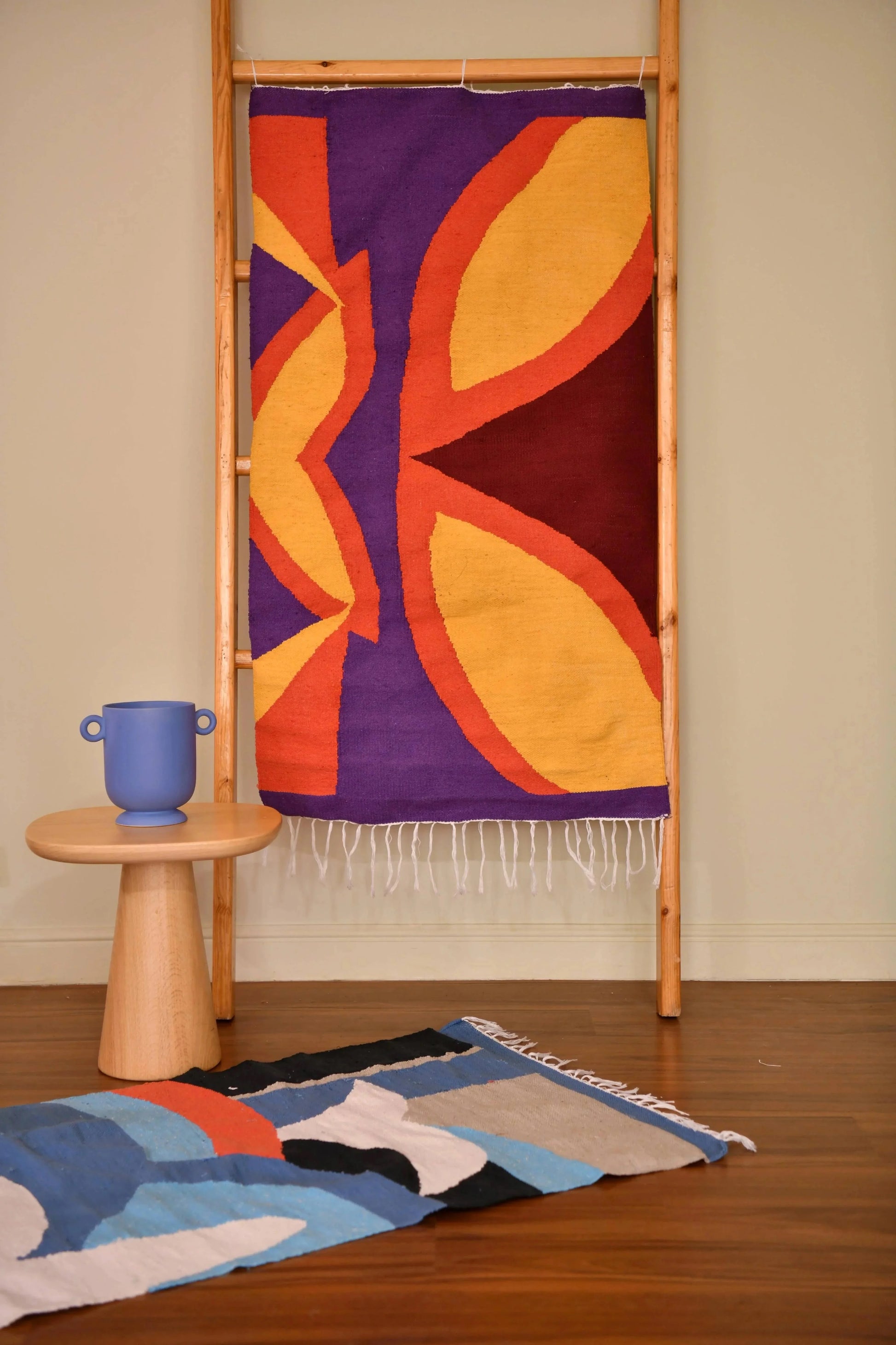 A colorful woven rug displayed on a wooden rack, featuring bold shapes in yellow, orange, and purple. Beside it, another smaller rug with a blue and black design is partially visible. A wooden cylindrical pedestal with a blue vase sits next to the rack.