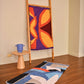 "A colorful textile hanging on a wooden ladder, featuring bold patterns in purple, orange, and yellow. To the left, a vibrant blue, black, and white rug lies on a wooden floor. A small, round wooden table with a blue vase sits next to the textile display."