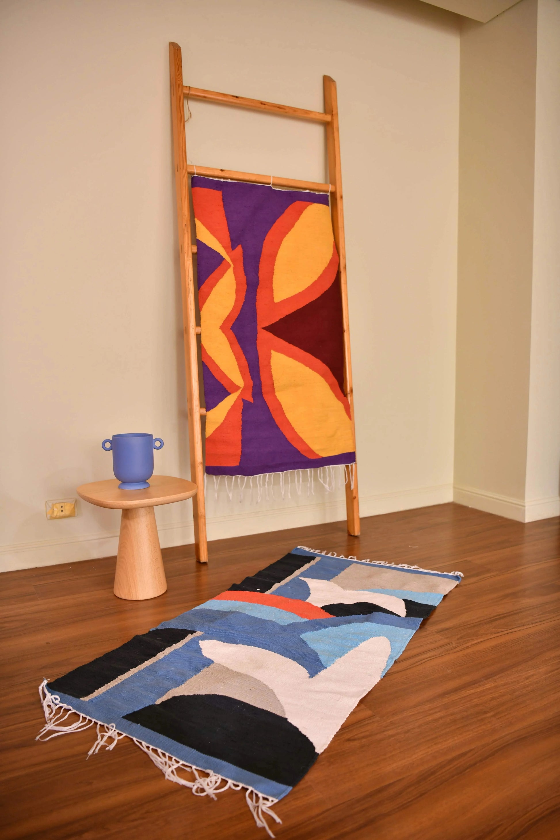 "A colorful textile hanging on a wooden ladder, featuring bold patterns in purple, orange, and yellow. To the left, a vibrant blue, black, and white rug lies on a wooden floor. A small, round wooden table with a blue vase sits next to the textile display."