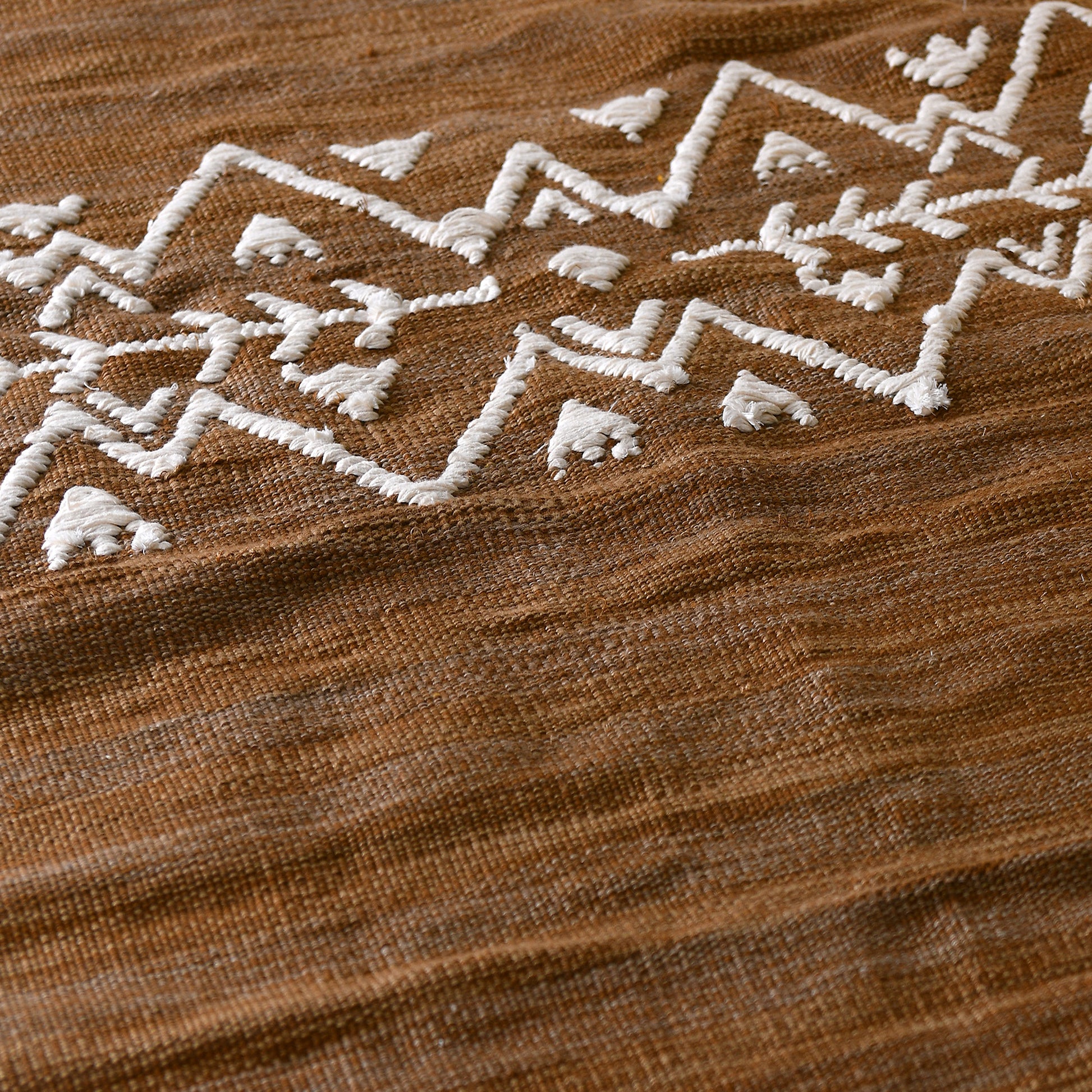 A long, narrow rug in a light brownish-tan color is shown against a white background. The rug has a slightly textured, woven appearance with subtle variations in shade. Near the bottom, a band of white geometric designs is woven into the rug. The pattern consists of symmetrical rows of white triangles, zigzags, and stylized arrow shapes, creating a visually appealing and slightly bohemian aesthetic. The overall style of the rug is simple yet elegant with a size of 1 x 2 meters.