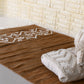 A long, narrow rug in a light brownish-tan color is shown against a white background. The rug has a slightly textured, woven appearance with subtle variations in shade. Near the bottom, a band of white geometric designs is woven into the rug. The pattern consists of symmetrical rows of white triangles, zigzags, and stylized arrow shapes, creating a visually appealing and slightly bohemian aesthetic. The overall style of the rug is simple yet elegant with a size of 1 x 2 meters.