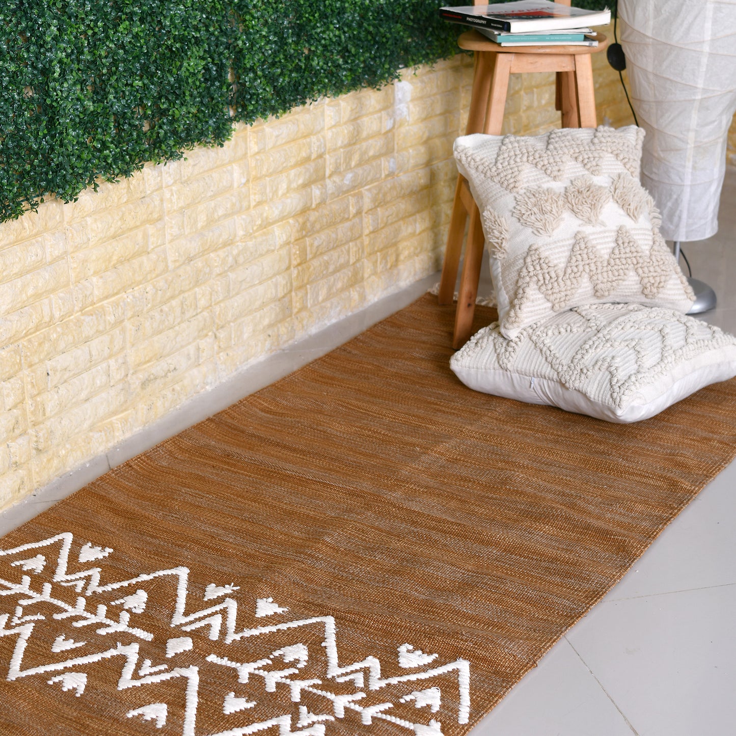 A long, narrow rug in a light brownish-tan color is shown against a white background. The rug has a slightly textured, woven appearance with subtle variations in shade. Near the bottom, a band of white geometric designs is woven into the rug. The pattern consists of symmetrical rows of white triangles, zigzags, and stylized arrow shapes, creating a visually appealing and slightly bohemian aesthetic. The overall style of the rug is simple yet elegant with a size of 1 x 2 meters.