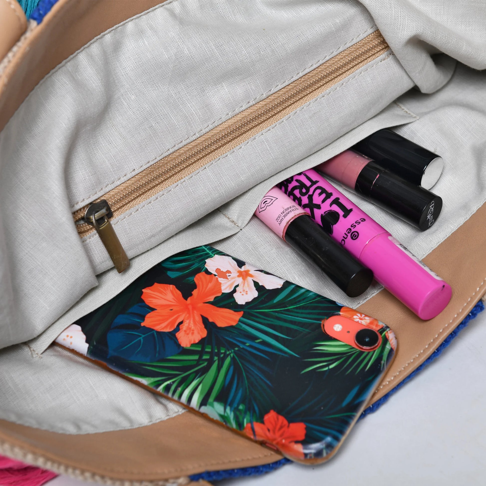 "An open handbag showing a light-colored interior with a zipper pocket. Inside, there are several cosmetics including lipsticks in black and pink tubes, and a phone case with a tropical floral design."