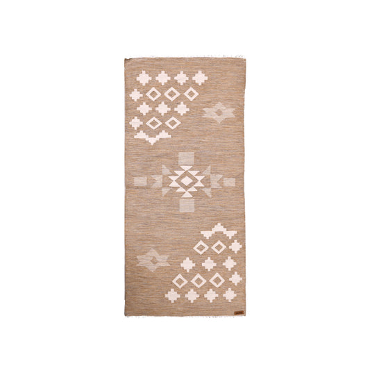 A rectangular woven rug is displayed against a white background. The rug features a neutral beige color with intricate white geometric patterns, including diamonds and crosses. The design combines modern and traditional elements, while the fringed edges add texture. A small brand tag is visible in the corner, enhancing the rug's stylish appeal.