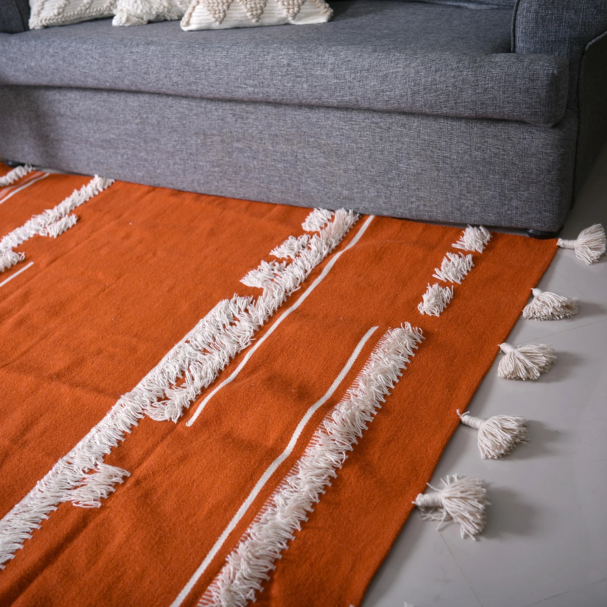 A close-up view of an orange area rug featuring white fringes and decorative stripes, positioned in front of a gray sofa. The rug's vibrant color and texture contrast beautifully with the smooth floor beneath it. The soft, inviting appearance adds warmth to the living space, enhancing the overall cozy atmosphere.