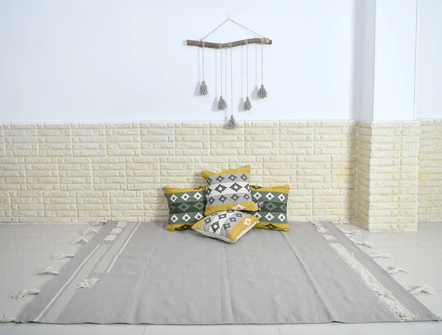 The image features a minimalist interior setting with a light-colored, textured wall and a smooth floor. In the center, there is a large, neutral-toned rug adorned with decorative edges. A collection of patterned cushions in green, yellow, and white are neatly arranged against the wall. Above the cushions, there is a bamboo wall hanging with tassels, adding a touch of bohemian style to the decor. The overall atmosphere is calm and inviting, emphasizing simplicity and comfort.