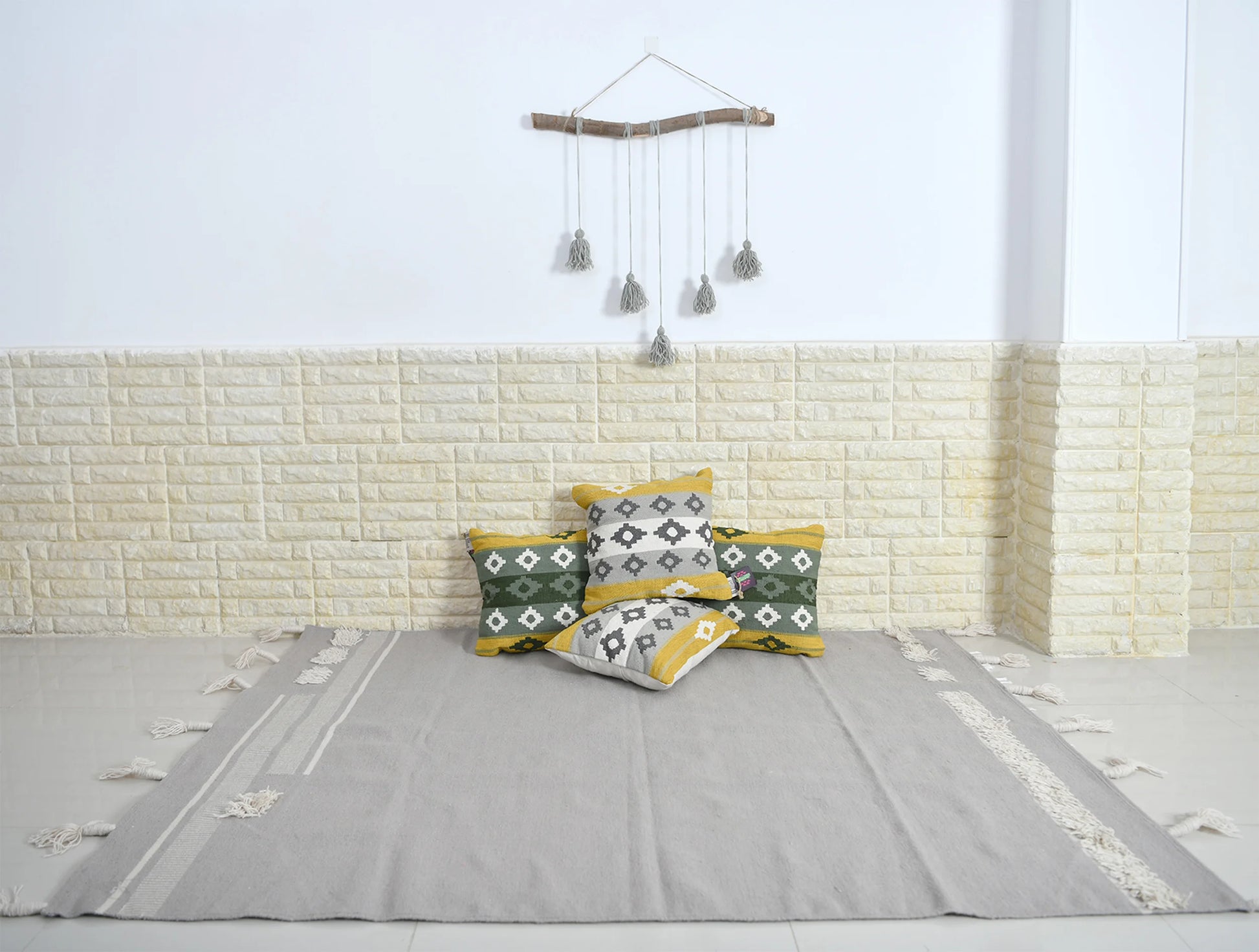 The image features a minimalist interior setting with a light-colored, textured wall and a smooth floor. In the center, there is a large, neutral-toned rug adorned with decorative edges. A collection of patterned cushions in green, yellow, and white are neatly arranged against the wall. Above the cushions, there is a bamboo wall hanging with tassels, adding a touch of bohemian style to the decor. The overall atmosphere is calm and inviting, emphasizing simplicity and comfort.