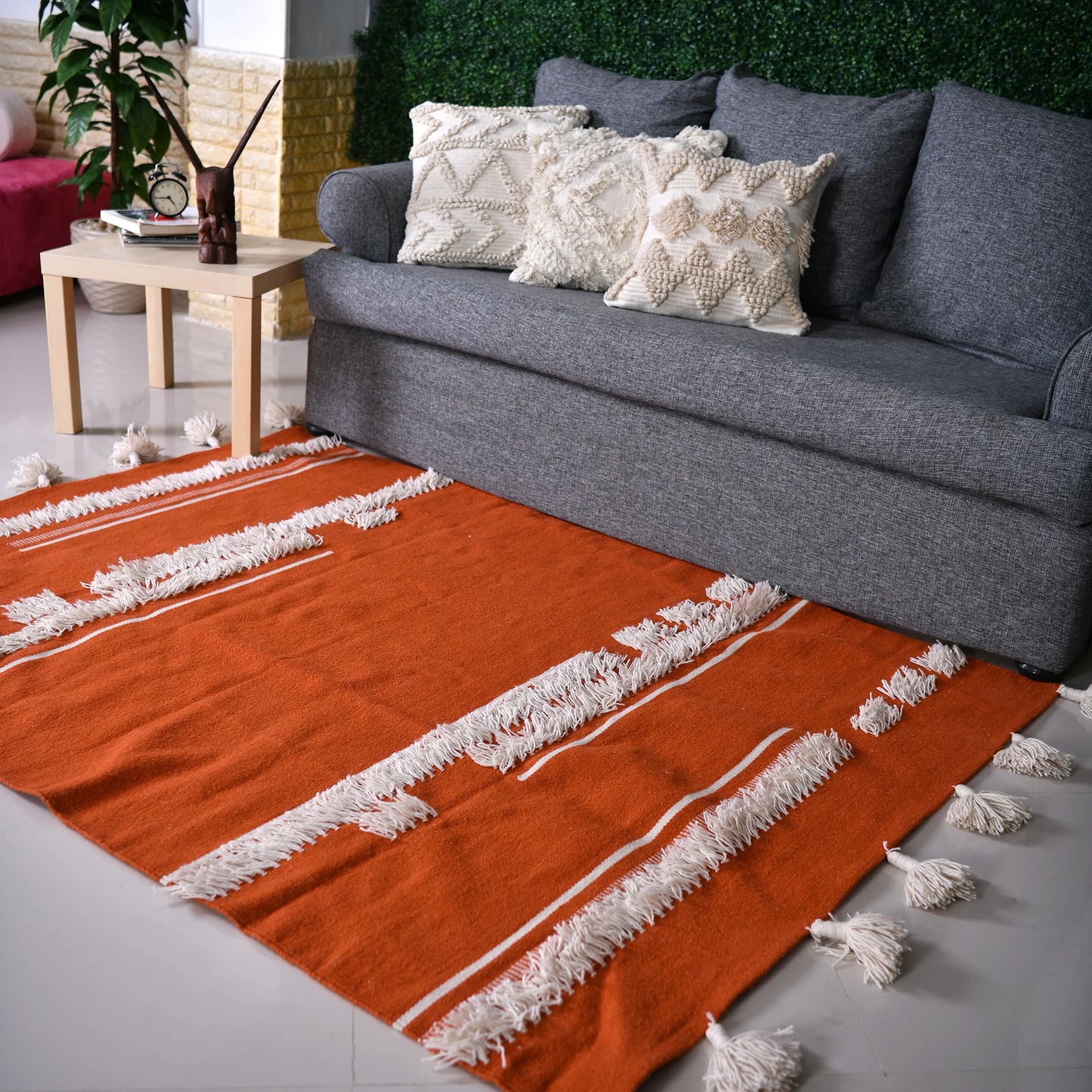 A stylish living room featuring a gray sofa adorned with decorative pillows in various textures. The sofa is positioned on an eye-catching orange area rug with white fringes. To the left, a light wood side table holds a decorative sculpture. The backdrop includes a vibrant green wall of faux foliage, enhancing the room's modern aesthetic. Natural light floods the space, creating a welcoming ambiance.