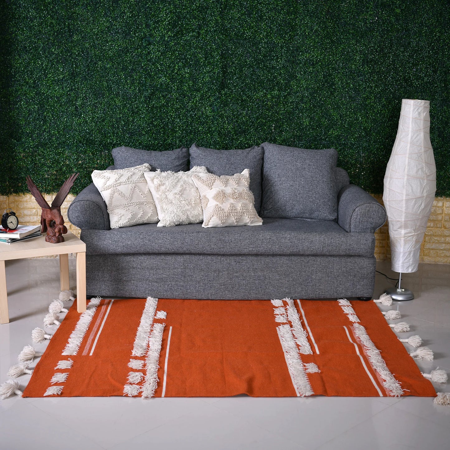 A modern living room setup featuring a gray sofa adorned with decorative pillows and placed on an orange area rug with white fringes. To the left, a light wood side table holds a sculptural piece and a clock. A stylish floor lamp stands beside the sofa, and the backdrop consists of a vibrant green wall made of faux foliage, adding a touch of nature to the space.