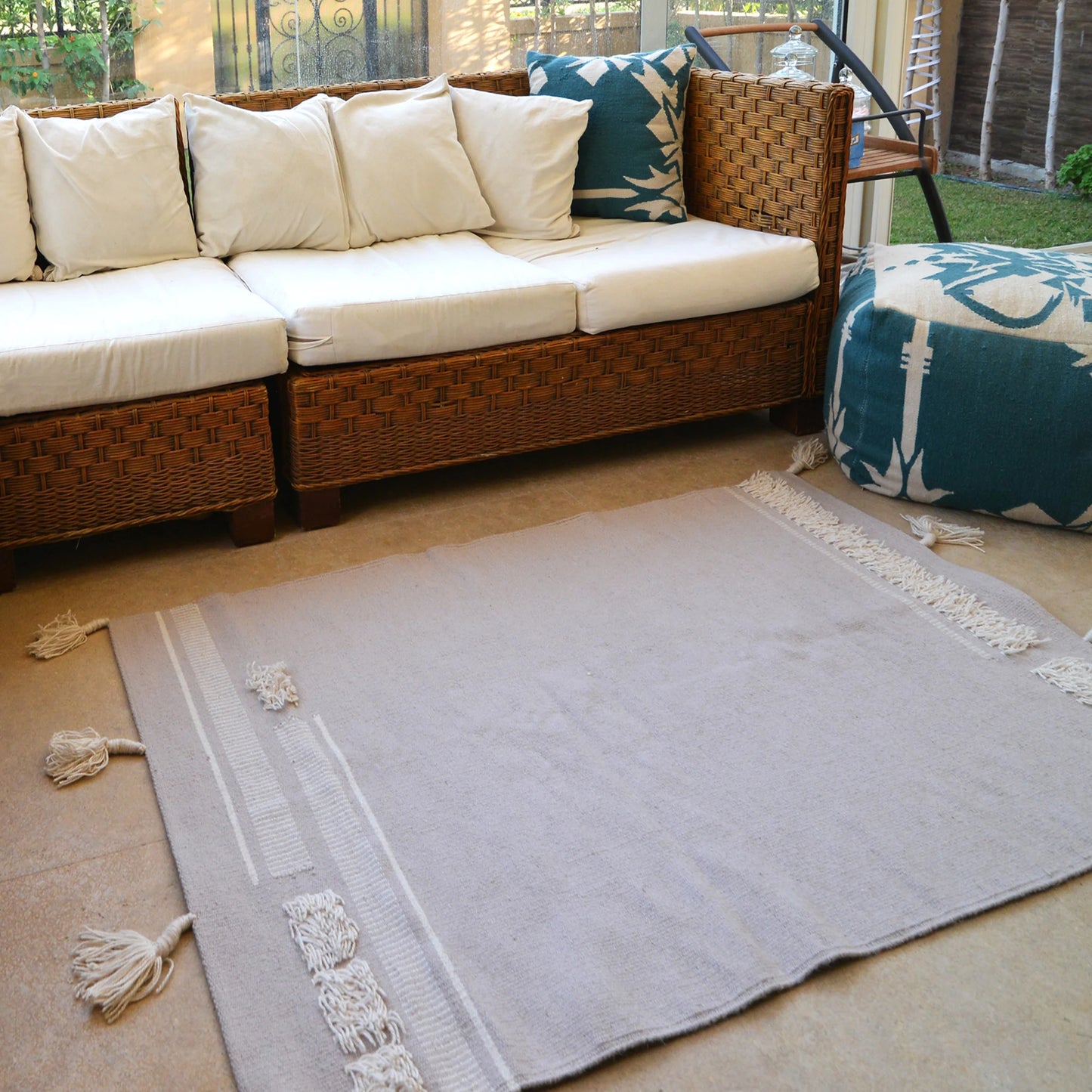 A cozy sunroom featuring a wicker sectional sofa with light beige cushions. In front of the sofa, a soft beige area rug with white fringes lies on the floor. To the right, a round ottoman in a bold teal and white pattern adds a decorative touch. Large windows allow natural light to illuminate the space, creating a warm and inviting atmosphere.