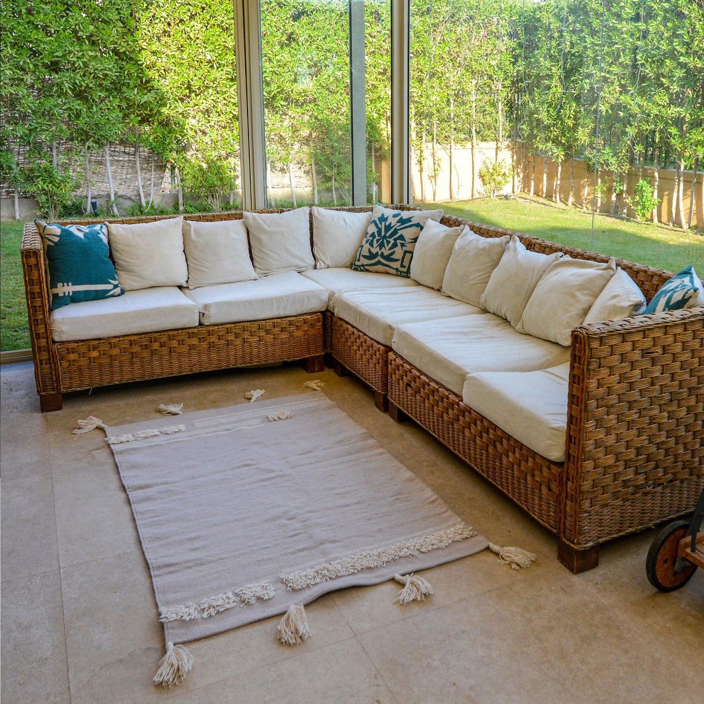 A bright and inviting sunroom featuring a large wicker sectional sofa with light beige cushions. In front of the sofa, a soft beige area rug with white fringes lies on the floor. Large glass windows showcase a lush green garden outside, creating a serene atmosphere. Decorative pillows in shades of teal and white add a pop of color to the sofa.