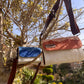 Two woven bags hang from a tree branch against a bright blue sky. The smaller bag features a blue and white design, while the larger bag displays a warm beige and terracotta pattern. Both bags have brown leather straps, creating a stylish contrast with the natural setting.