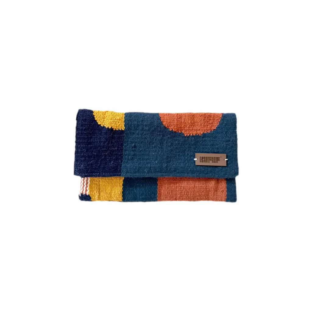 This image shows a clutch. The clutch has a geometric pattern in shades of navy blue, mustard yellow, and burnt orange. The front of the clutch has a small leather logo tag on it. The clutch appears to be made of a plush, textured material, likely yarn or thread. The image is against a plain white background, highlighting the vibrant colors and pattern of the unique handmade accessory.