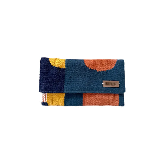 This image shows a clutch. The clutch has a geometric pattern in shades of navy blue, mustard yellow, and burnt orange. The front of the clutch has a small leather logo tag on it. The clutch appears to be made of a plush, textured material, likely yarn or thread. The image is against a plain white background, highlighting the vibrant colors and pattern of the unique handmade accessory.