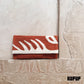 "A woven clutch in terracotta with abstract white leaf patterns, placed against a textured stone background featuring ancient hieroglyphics. The design blends modern aesthetics with historical elements."