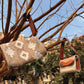 Two stylish handbags are hanging from the branches of a tree against a clear blue sky. The larger bag has a woven design featuring geometric patterns in earthy tones, while the smaller bag has a metallic finish with a textured strap. The natural setting highlights the bags' unique craftsmanship, with greenery visible in the background.