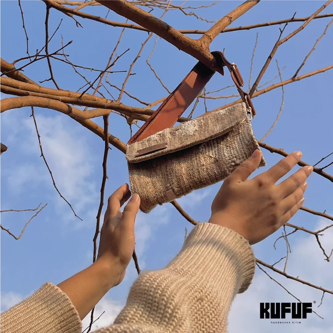 A person is holding a stylish handbag against a backdrop of a clear blue sky, with branches of a tree visible. The handbag features a textured woven design in neutral tones and has a brown leather strap. The brand name "KUFUF" is displayed in the bottom corner. The cozy, casual vibe is enhanced by the person's light-colored sweater, creating a warm and inviting scene.