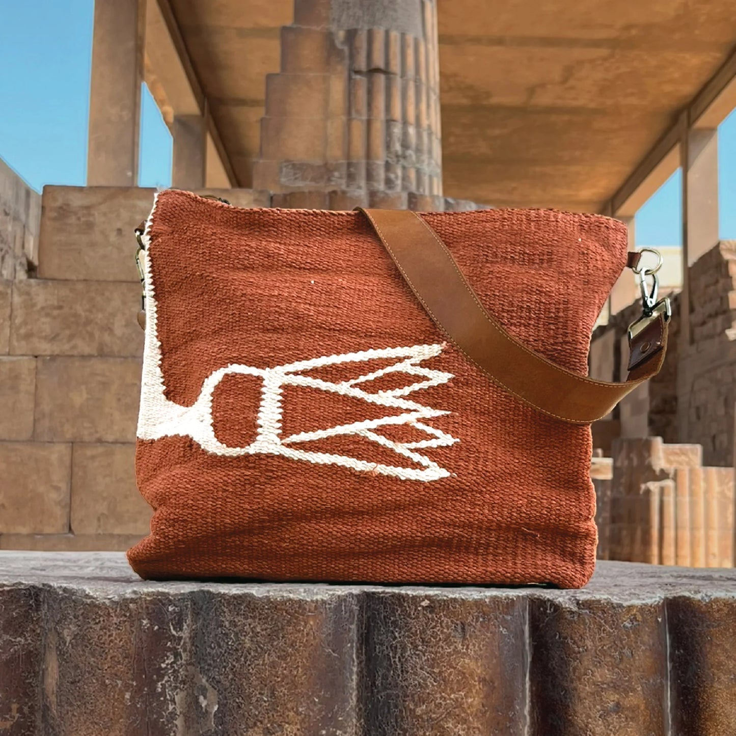 "A terracotta woven shoulder bag with a bold white abstract design resembling a leaf or flower, positioned on a textured stone surface. The bag features a brown leather strap and is set against a backdrop of ancient architectural columns."