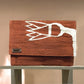 A rectangular clutch bag is showcased on a wooden shelf against a soft, neutral background. The bag is in a terracotta hue with a bold white woven floral pattern. The metal brand tag "KUFUF" is visible, emphasizing the bag's stylish and contemporary design. The overall presentation highlights its artistic and modern appeal.