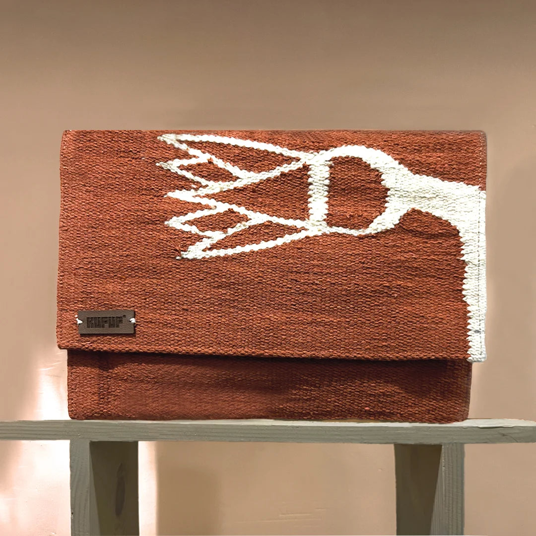 A rectangular clutch bag is showcased on a wooden shelf against a soft, neutral background. The bag is in a terracotta hue with a bold white woven floral pattern. The metal brand tag "KUFUF" is visible, emphasizing the bag's stylish and contemporary design. The overall presentation highlights its artistic and modern appeal.