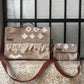 Two woven bags are displayed against a grid-patterned window. The larger bag features a warm beige background with white geometric patterns and fringed detailing at the bottom, complemented by a brown leather strap. Next to it, a smaller bag shares a similar design aesthetic, with the same patterns and a leather strap. The textured surfaces of the bags create a stylish and cohesive look.