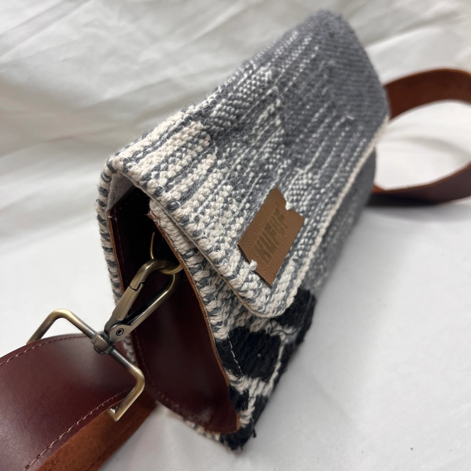 "A side view of a gray woven bag with a textured pattern and a brown leather strap. The image captures the bag's details, including the leather tag that reads 'KUFUF.'"