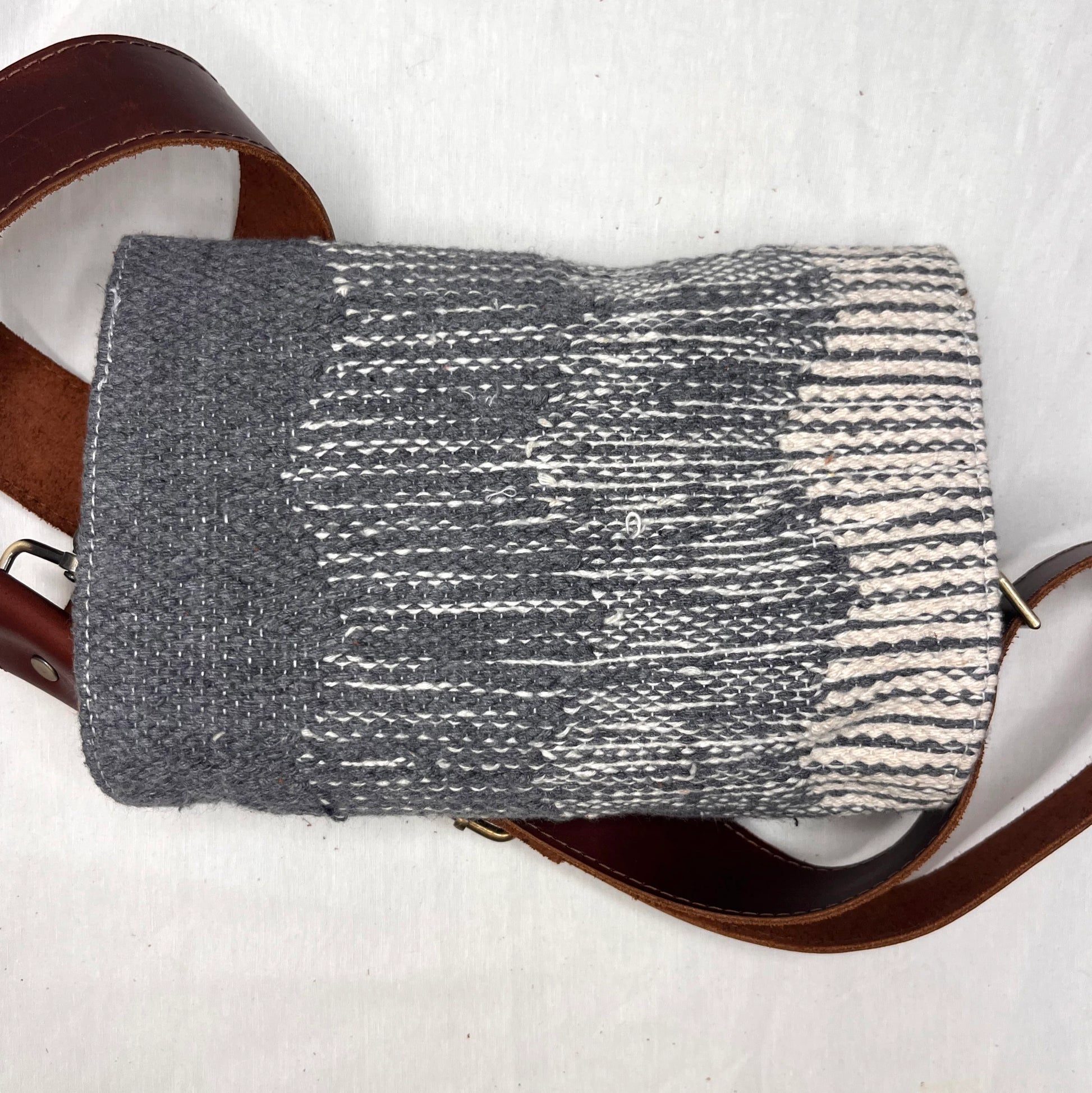 "A top view of a gray woven bag with a striped pattern and a brown leather strap. The bag is resting on a white surface, emphasizing its design and texture."