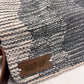 "Close-up of a gray woven bag displaying its textured pattern, with a leather tag embossed with 'KUFUF.' The image highlights the intricate craftsmanship of the material."