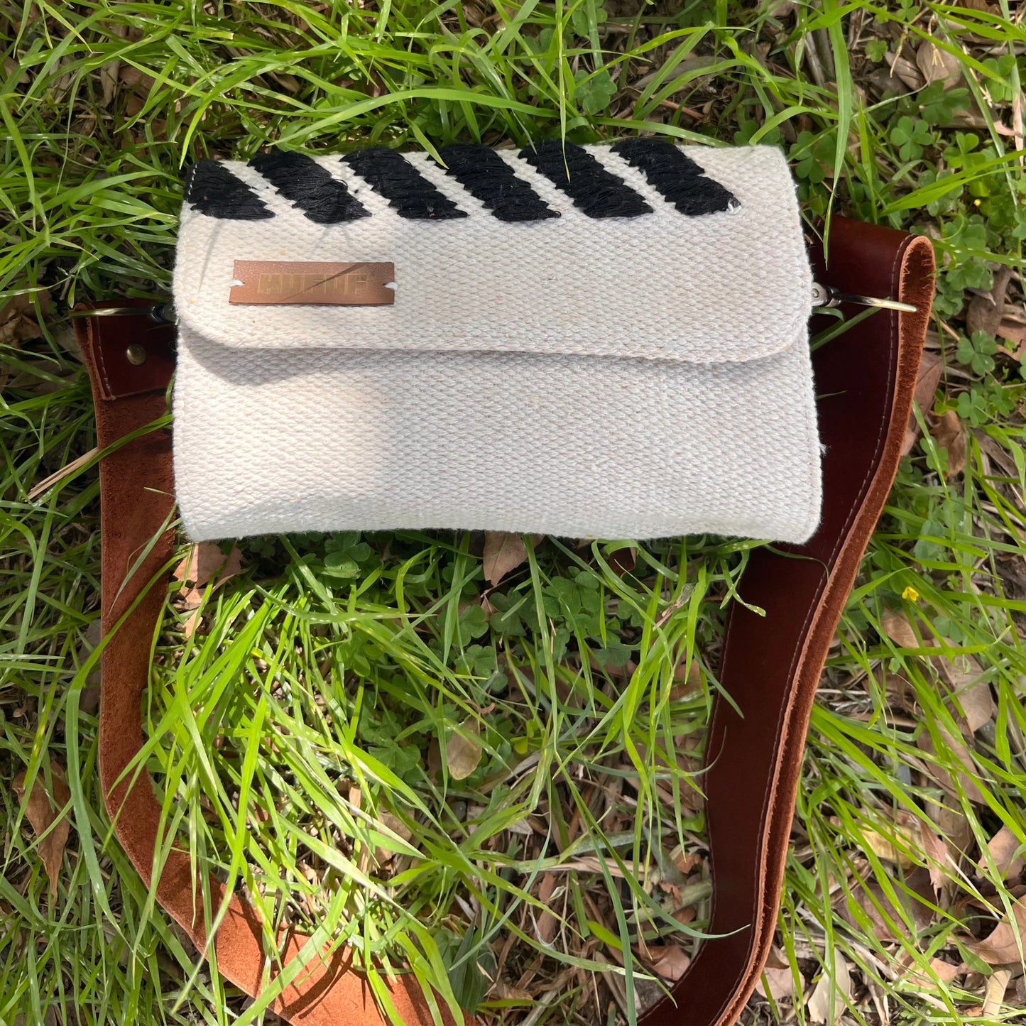 "A cream-colored woven bag with black stripes, resting on green grass. The bag has a brown leather strap and a metal tag embossed with 'KUFUF,' blending contemporary design with a natural setting."