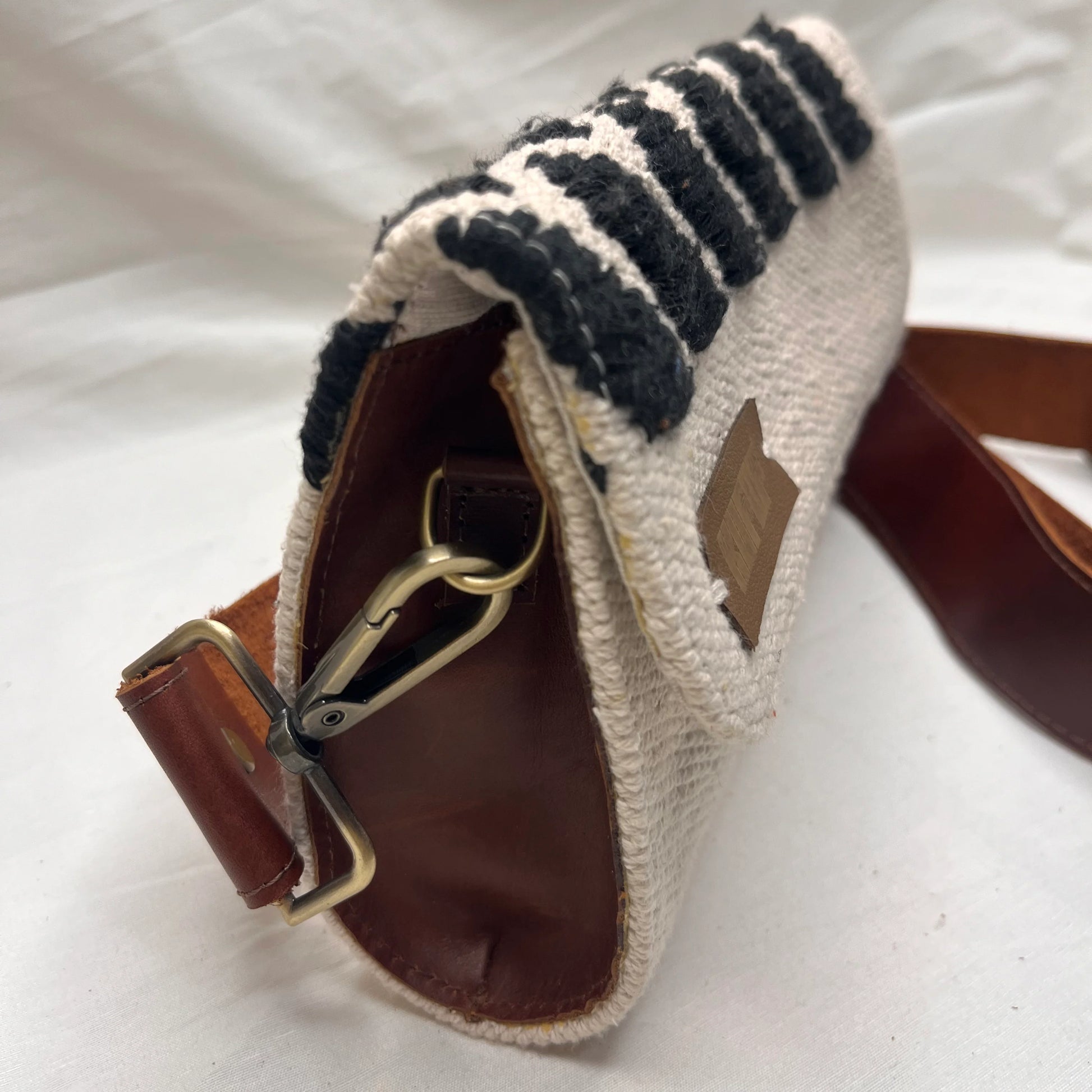 "A close-up of a cream-colored woven bag featuring black stripes and a brown leather strap. The image captures the bag's side view, showcasing its clasp and leather tag."